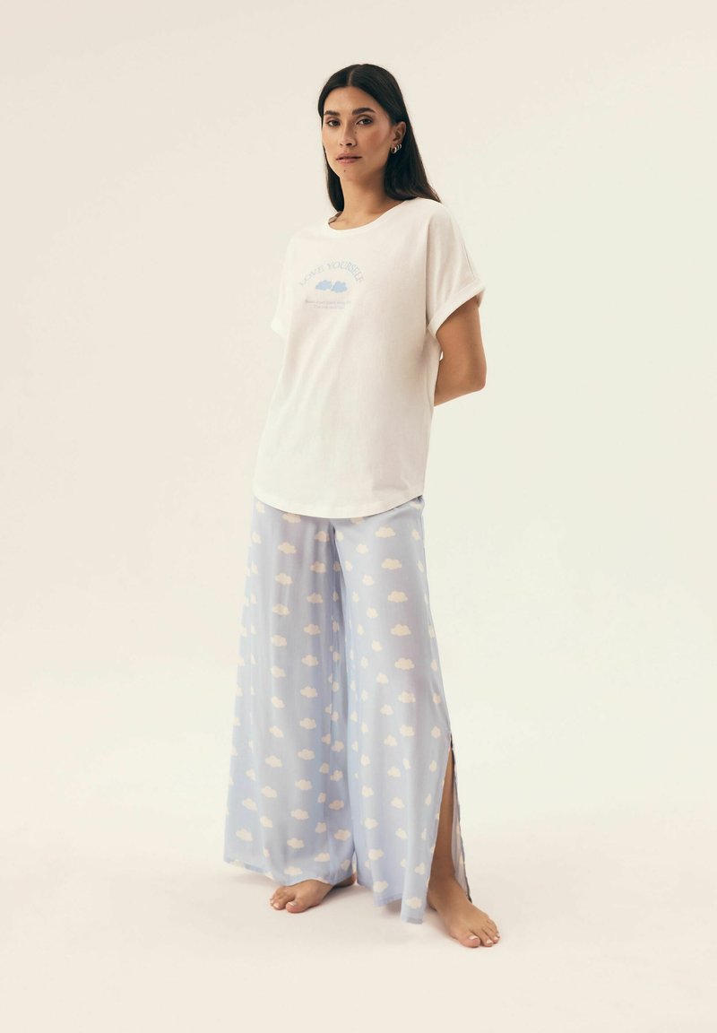 Biała bawełniana koszulka z nadrukiem "LOVE YOURSELF" w jasnoniebieskim kolorze; zestawiona z jasnoniebieskimi szerokimi spodniami z białymi wzorami chmur i bocznymi rozcięciami.