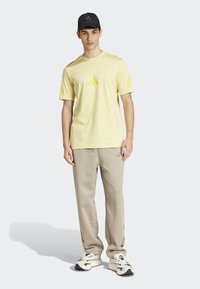 Žluté tričko s krátkým rukávem a třemi diagonálními pruhy na hrudi, spárované s béžovými kalhotami a černými teniskami.