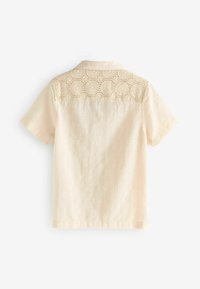 Kurze Bluse aus hellbeigem Stoff, mit strukturiertem Grund und Spitzenpaneel mit kreisförmigen Mustern auf der Rückseite.