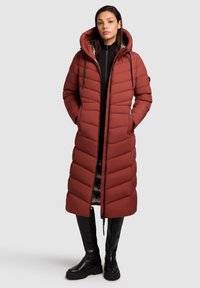 Long, rust-colored puffer coat with chevron stitching, a drawstring hood, front zipper, and side pockets. Worn over black leggings and boots.
