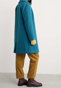 Blågrøn uldfrakke med krave og sideslidser; båret over en gul sweater og kombineret med sennepsfarvede fløjlsbukser og bordeaux støvler.