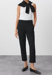 Svarta skräddarsydda byxor med smal passform och ankellängd, tillsammans med en vit linne och en svart scarf knuten runt halsen.