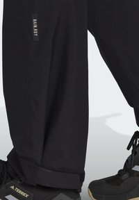 Pantaloni neri con un design leggermente affusolato, dotati di una piccola etichetta con logo e orlo a risvolto, abbinati a scarpe da ginnastica nere.