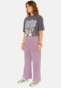 T-shirt graphique gris avec un design AC/DC ; associé à un pantalon en velours côtelé large de couleur lilas clair ; modèle portant des baskets argentées.