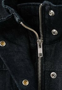 Schwarze Jeansjacke mit einem silbernen Reißverschluss und messingfarbenen Druckknöpfen, aus strukturiertem Stoff und mit einem umgeschlagenen Kragen, der detaillierte Nähte zeigt.