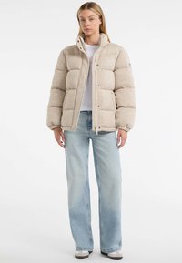 Jachetă puffer bej cu guler înalt și fermoar pe față, cu un design matlasat și manșete ribbate, asortată cu blugi albaștri deschiși.