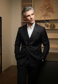 Sort dobbeltbrystet jakkesæt med en tekstureret overflade, parret med en hvid skjorte. Baggrunden har neutrale nuancer og moderne reoler.