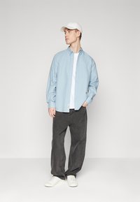 Chemise bleu clair à manches longues, associée à un t-shirt blanc à col rond et un pantalon large gris foncé. Baskets blanches visibles.
