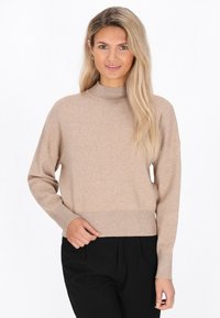 Beige gestrickter Pullover mit hohem Stehkragen, abgesenkten Schultern und geripptem Saum. Kombiniert mit schwarzen Hosen für einen Kontrast. Weiche Textur.