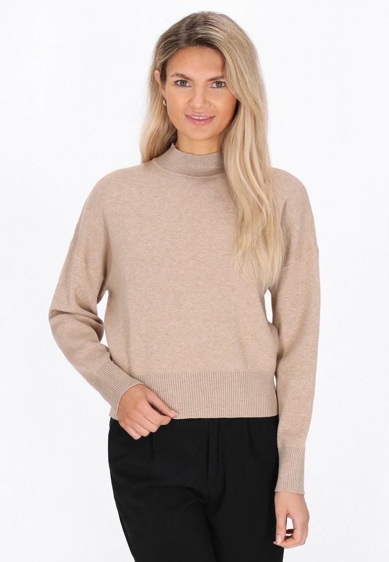 Beige gestrickter Pullover mit hohem Stehkragen, abgesenkten Schultern und geripptem Saum. Kombiniert mit schwarzen Hosen für einen Kontrast. Weiche Textur.