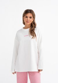 Witte blouse met lange mouwen en een ronde hals, voorzien van een roze "MIRONS" logo op de borst; gecombineerd met roze gestreepte broek.