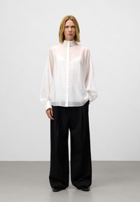 Witte, doorzichtige blouse met lange mouwen en hoge hals, gecombineerd met zwarte wijde broek. De blouse heeft een knoopsluiting aan de voorkant en een doorzichtige textuur.