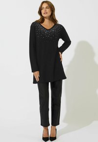 Schwarzer Pullover mit V-Ausschnitt und langen Ärmeln, verziert mit silbernen Akzenten entlang des Ausschnitts. Kombiniert mit schwarzen Hosen und Heels.