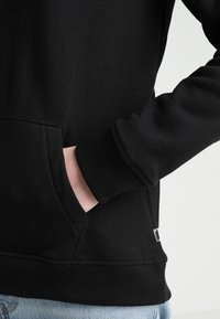 Czarna bluza z kapturem, z kieszenią kangurzą, teksturowana tkanina, ściągacze przy mankietach i u dołu, oraz mała metka marki z boku.