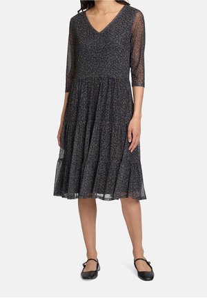 Femme portant une robe noire mi-longue avec des manches 3/4 transparentes et de subtils pois gris, accompagnée de ballerines Mary Jane noires.