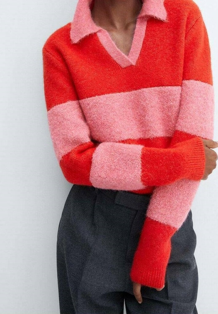 Persoon in een rode en roze gestreepte fuzzy trui met een kraag, gecombineerd met hooggetailleerde donkergrijze broek, met de armen gekruist op taillehoogte.