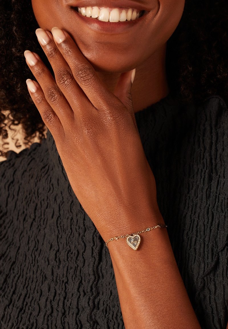 Gouden armband in de vorm van een hart met een textuur en een schakelkettinkje ontwerp. Gekenmerkt op een bruine hand tegen een donkere stoffen achtergrond.