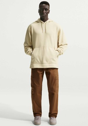 Homme debout, les mains dans les poches, portant un hoodie beige clair, un pantalon cargo marron et des baskets claires, sur un fond uni.