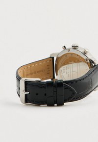 Ασημένιο ρολόι χειρός με μαύρη δερμάτινη αλυσίδα, μεταλλικό κούμπωμα και ορατό χαραγμένο πίσω μέρος που δείχνει λεπτομέρειες της μάρκας και του μοντέλου.