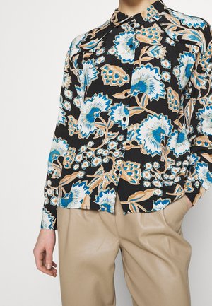 Zwart overhemd met knopen en grote bloemenprint in blauw, beige en wit, gedragen met beige broek, hand ontspannen langs het lichaam.