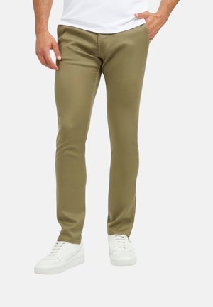 Uomo che indossa pantaloni kaki slim fit e sneakers bianche, in piedi con una mano in tasca, camicia bianca parzialmente visibile.