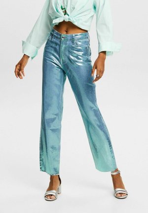 Frau trägt glänzende türkisfarbene High-Waist-Straight-Leg-Jeans, ein kurz geschnittenes hellgrünes, gebundenes Hemd und silberne Sandalen mit Absatz.