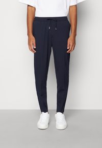 Man som bär marinblå drachansbyxor och vita sneakers, står mot en enkel ljus bakgrund.