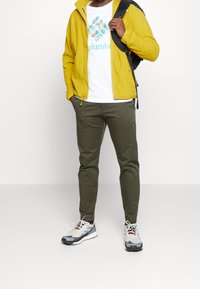 Gul zip-up jacka, vit t-shirt med turkos logotyp, olivgröna byxor och grå sneakers med orange accenter. Strukturerade material.