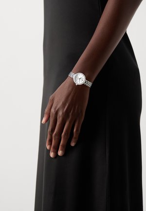 Silberne Armbanduhr mit einem weißen Ziffernblatt, verlinktem Armband und einem runden Gehäuse, getragen an einer Hand in einem dunkelstofflichen Kleid.