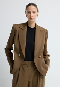 Ruskea kaksirivinen bleiseri, jossa on korostetut kaulukset ja kultasävyiset napit, yhdistettynä mustaan yläosaan ja samansävyisiin pleatattuihin housuihin. Sujuva tekstuuri.