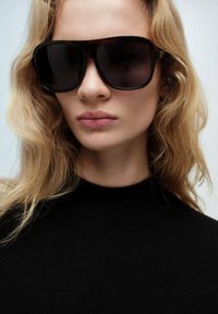 Occhiali da sole neri oversize con una montatura spessa e lenti scure. Il modello ha i capelli biondi ondulati e indossa un top nero con collo alto.