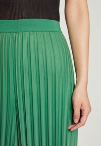 Detailaufnahme einer Person, die einen grünen, gefalteten Rock und ein schwarzes, geripptes Oberteil trägt, mit ihrem rechten Arm entspannt an ihrer Seite.