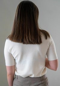 Persoon met stijlvol bruin haar in een witte trui met korte mouwen en grijze broek, weergegeven vanuit de rug tegen een effen achtergrond.