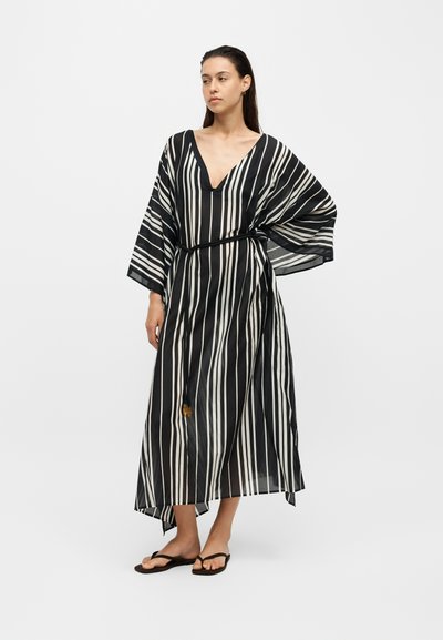 Femme debout portant une robe rayée verticale noir et blanc avec un décolleté en V profond et des manches larges, associée à des tongs noires.