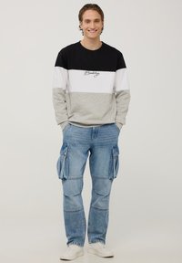 Męska bluza w kolorach czarnym, białym i szarym z napisem "Brooklyn"; w zestawie z jasnoniebieskimi spodniami cargo z wieloma kieszeniami. Białe sneakersy.