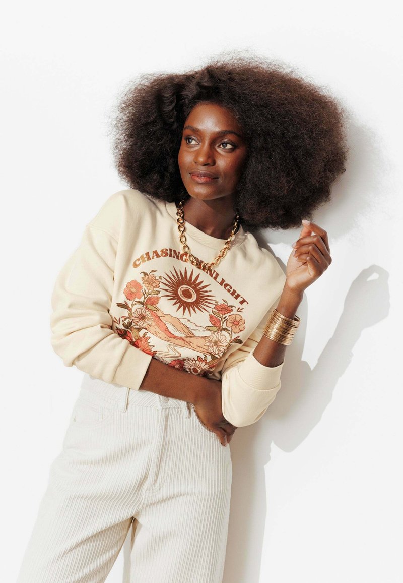 Femme aux cheveux bouclés volumineux portant un sweat-shirt crème avec un motif floral de soleil et le texte « Chasing the Sunlight », un collier en chaîne dorée et des bracelets.