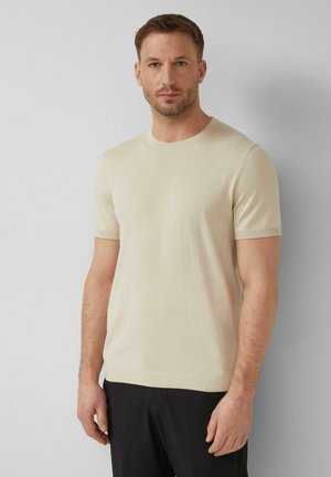 Mann mit kurzen braunen Haaren, der einen beigen kurzärmligen Pullover und schwarze Hosen trägt und vor einem einfarbigen grauen Hintergrund steht.