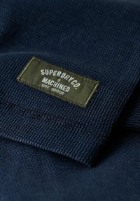 Navy blue knitted fabric with a green sewn-on label reading "SUPERDRY CO. MACHINED DRY GOODS."