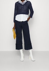 Marinblå kortstickad tröja med öppen väv, tillsammans med en vit skjorta med krage. Mörk marinblå vida byxor med volangdetaljer, vita blockhälsboots.