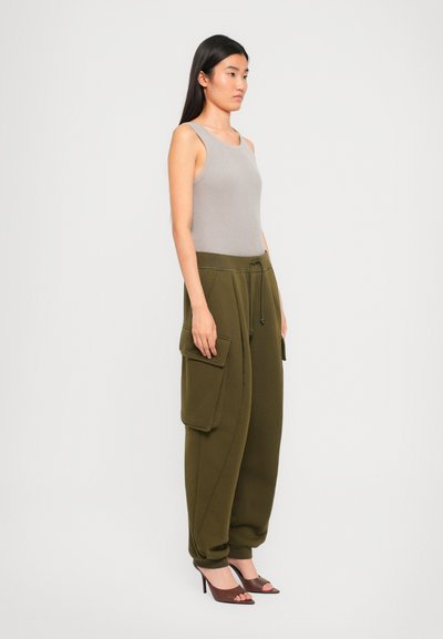 Woman standing in beige sleeveless ribbed top, olive green cargo pants with drawstring waist, and brown open-toe heels against a plain white background.