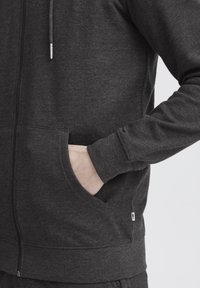 Donkergrijze zip-up hoodie met een gladde textuur, verstelbare trekkoorden, zijzakken en geribbelde boorden. Bevat een klein logolabel aan de onderkant.