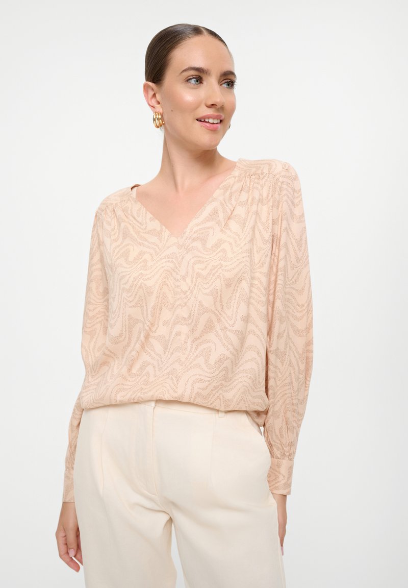 Blusa de cuello en V en color durazno claro con un patrón texturizado en forma de olas. Mangas largas con detalles gathered en los hombros, combinada con pantalones crema.