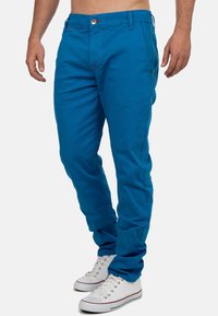 Man in blauwe slim-fit broek en witte sneakers, staand voor een effen witte achtergrond.