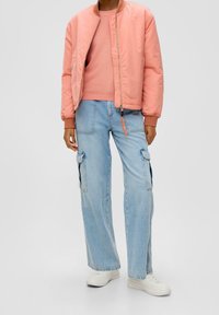 Veste bomber rose clair avec poignets côtelés portée sur un pull assorti, et jean cargo large bleu clair avec poches latérales, assortie à des baskets blanches.