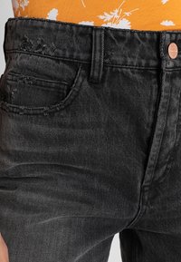 Pantalones vaqueros de mezclilla negra con un acabado texturizado, que presentan un diseño desgastado, estilo de cinco bolsillos y un cierre de botón metálico en la cintura.