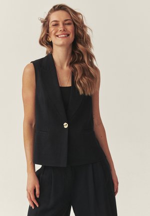 Šypsanti moteris su banguotais šviesiais plaukais, vilkinti be rankovių juodą švarką ir derančias kelnes, stovinti priešais šviesią, vientisą foną.