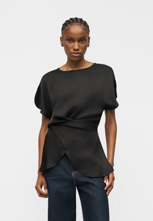 Femme aux cheveux tressés portant un chemisier noir à manches courtes et un jean bleu foncé, debout devant un fond clair uni.