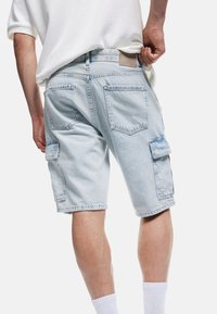 Pantalones cortos de mezclilla azul claro con un ajuste holgado, que cuentan con múltiples bolsillos, dobladillos deshilachados y un acabado texturizado. La parte trasera de la cintura tiene un parche de tela.