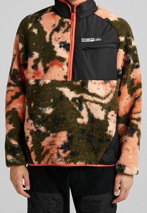 Fleecejacke in Grün, Pfirsich, schwarzem Camouflage-Muster mit schwarzer Brusttasche und orangefarbenem Reißverschlussbesatz, getragen von einer stehenden Person.