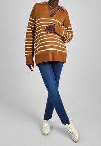Maglione marrone a righe con scollo a V e colletto, abbinato a jeans slim blu e sneakers bianche, esposto su uno sfondo chiaro.